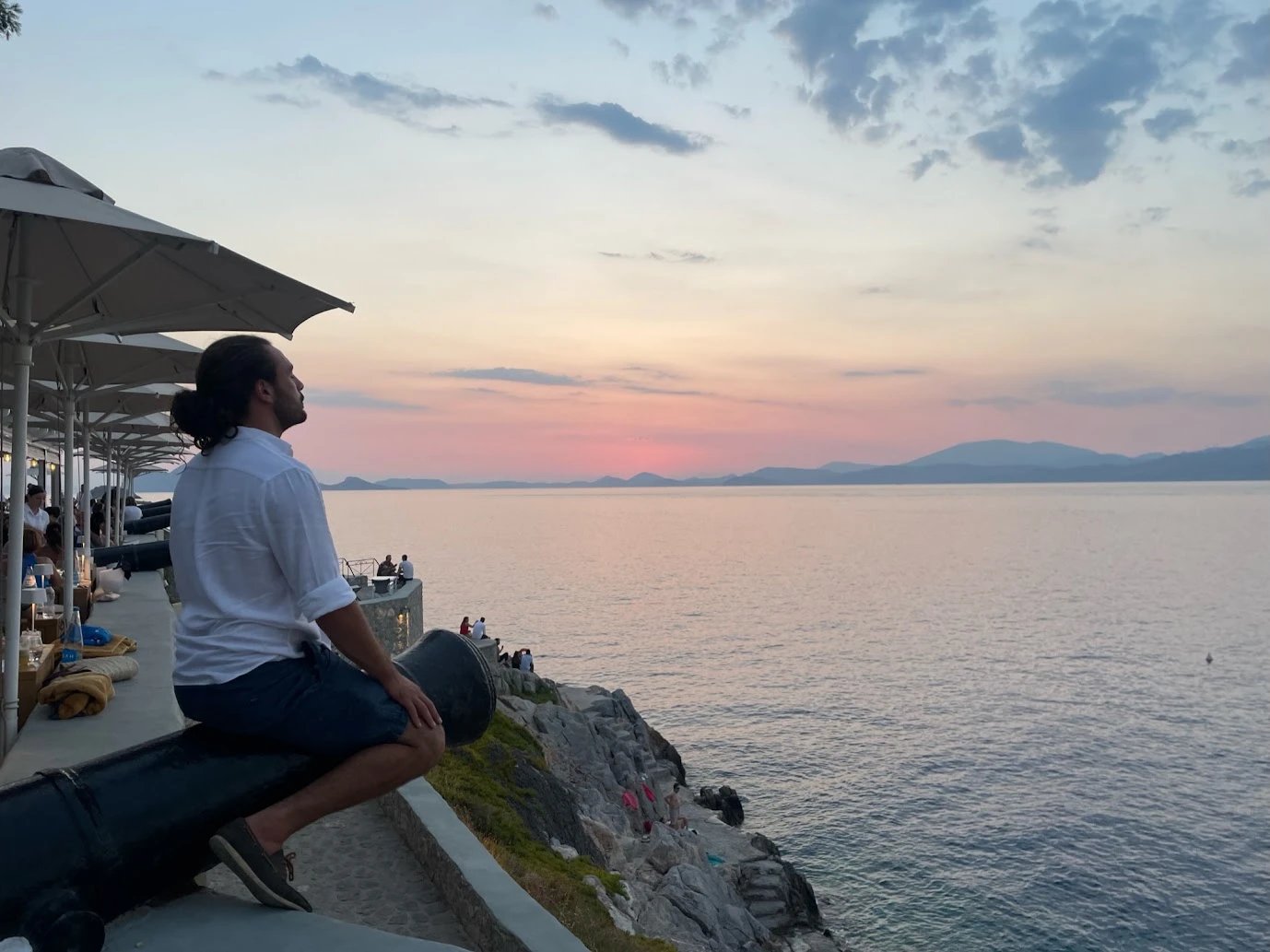 A man sitting on an old cannon on a seaside terrace, looking out at a pink sunset over the sea and distant mountains
