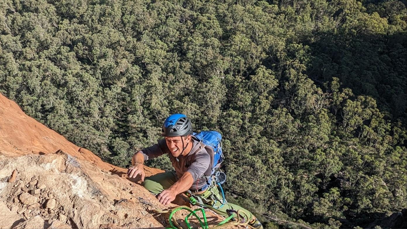 A climber in a blue helmet pulling over an orange sandstone edge, green rope below, forest far beneath